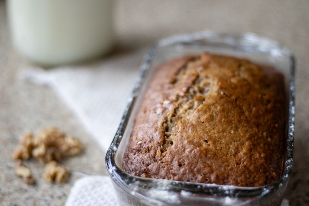 A loaf of baked sourdough banana nut bread in a glass loaf pan with nuts and milk in the background.