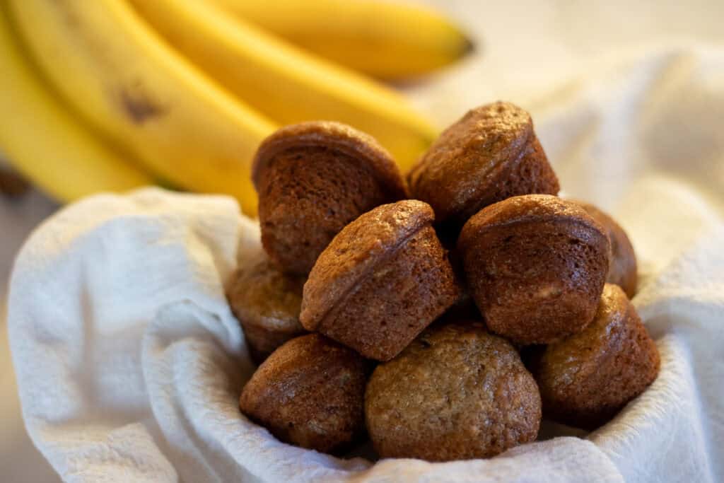 A tea-towel lined bowl filled with sourdough banana mini muffins and some bananas in the background.