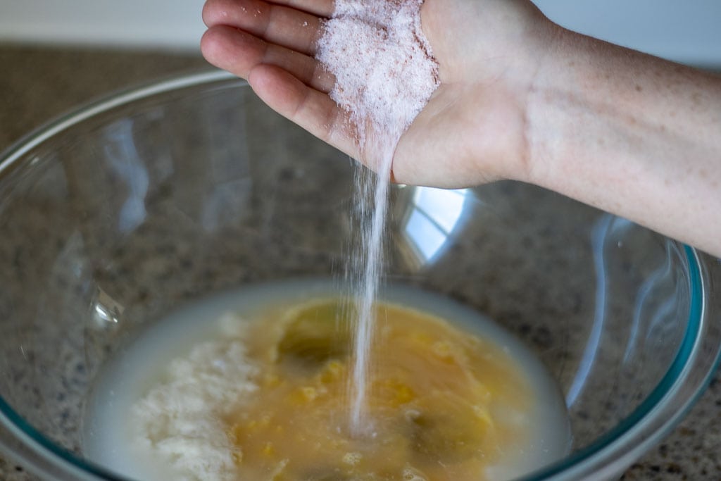A hand pouring sea salt into a bowl of bread dough ingredients.