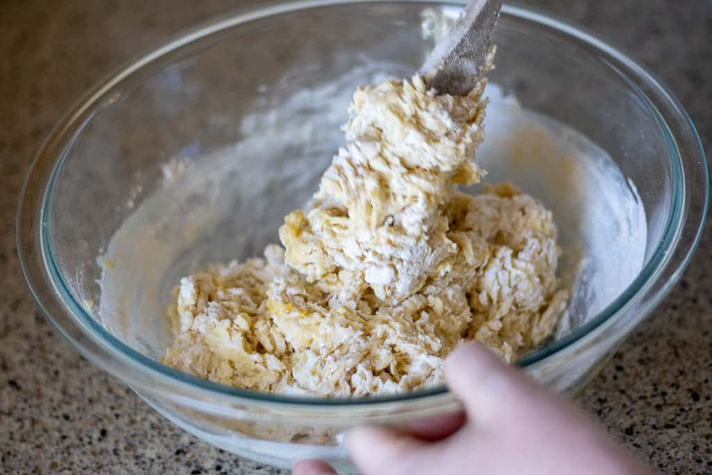 A hand using a wooden spoon to mix bread dough ingredients together.