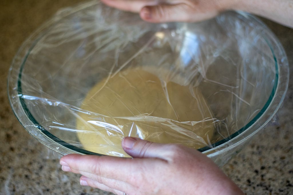 Hands using plastic wrap to cover a bowl of sourdough challah bread dough.