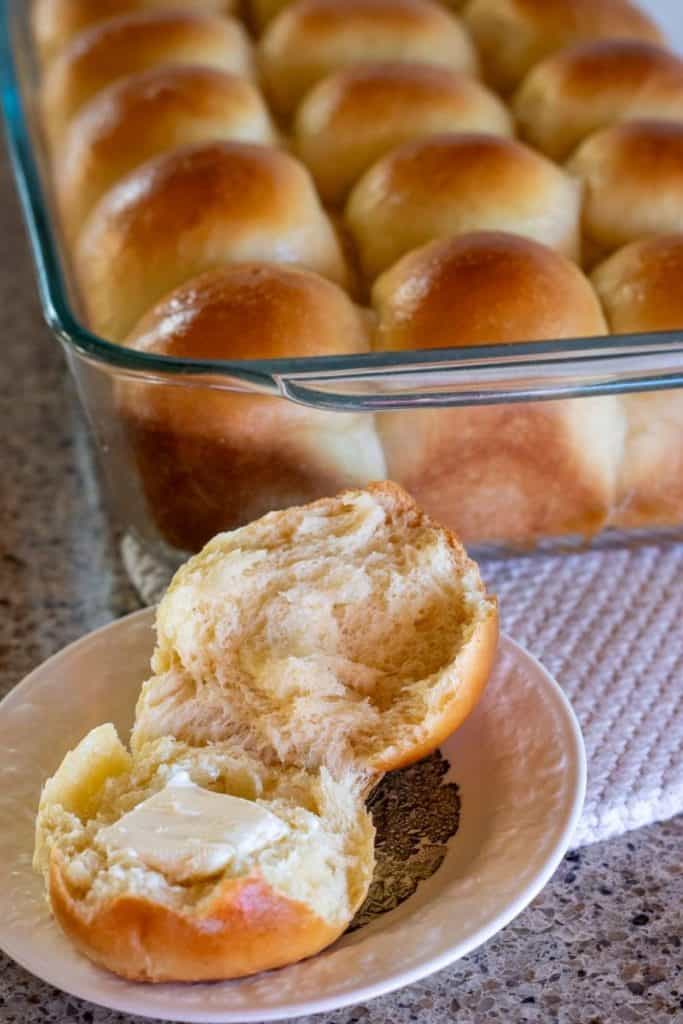 A buttered sourdough roll on a small plate next to a full pan of rolls.