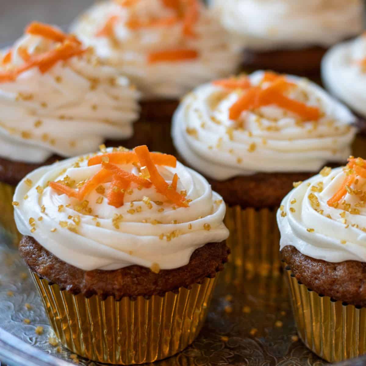 A batch of sourdough carrot cake muffins in golden cupcake liners topped with cream cheese frosting and shredded carrots.