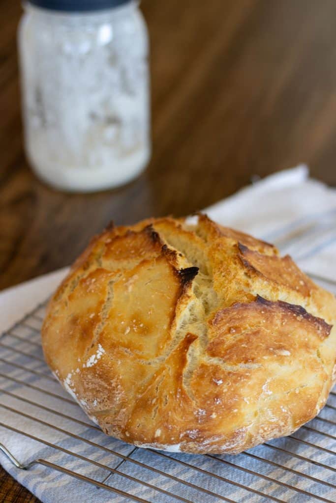 A loaf of sourdough bread set on a wire rack with a jar of sourdough starter in the background.