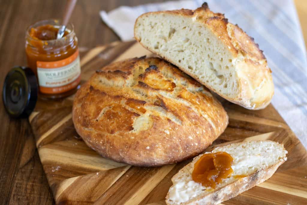 Two loaves of no stretch and fold sourdough bread set on a cutting board with a slice of bread with butter and jam.