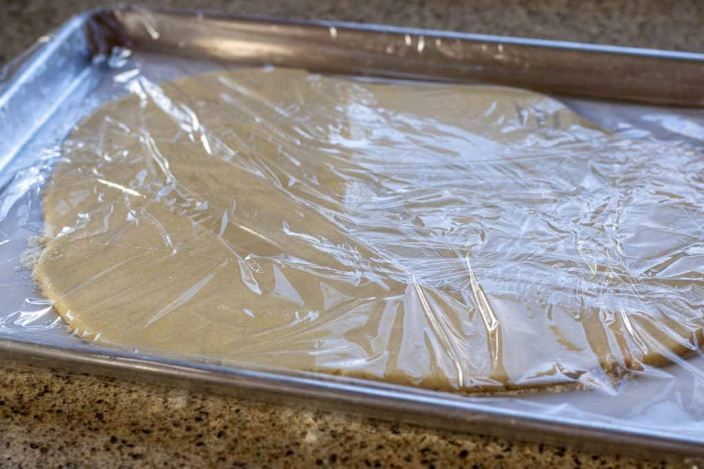 Shortbread cookie dough rolled out flat on a piece of parchment paper placed on a cookie sheet and covered with plastic wrap.