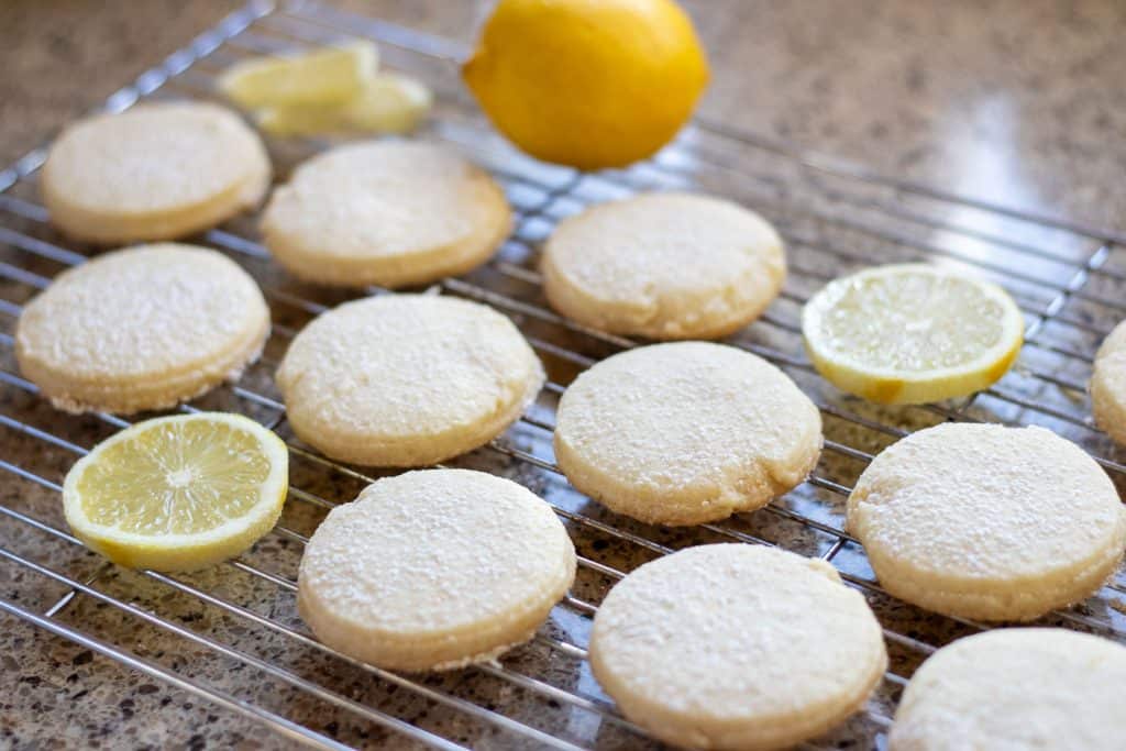 A batch of lemon shortbread cookies on a wire rake with a slice of lemon.