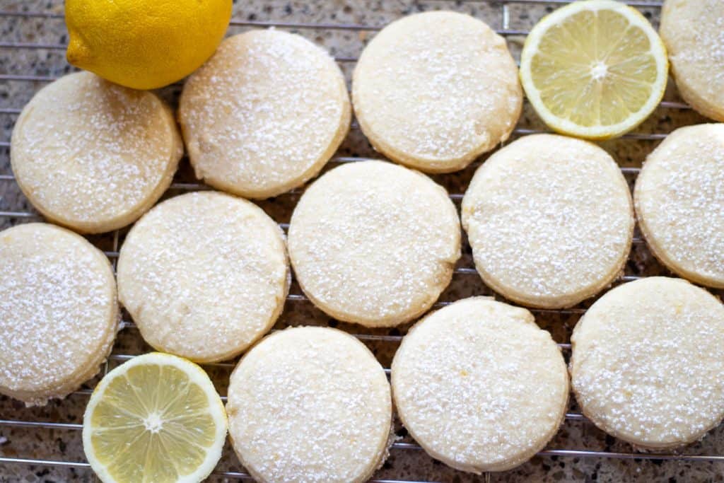 A batch of lemon-flavored shortbread cookies, dusted with powdered sugar, arranged on a wire rack with a lemon wedge.