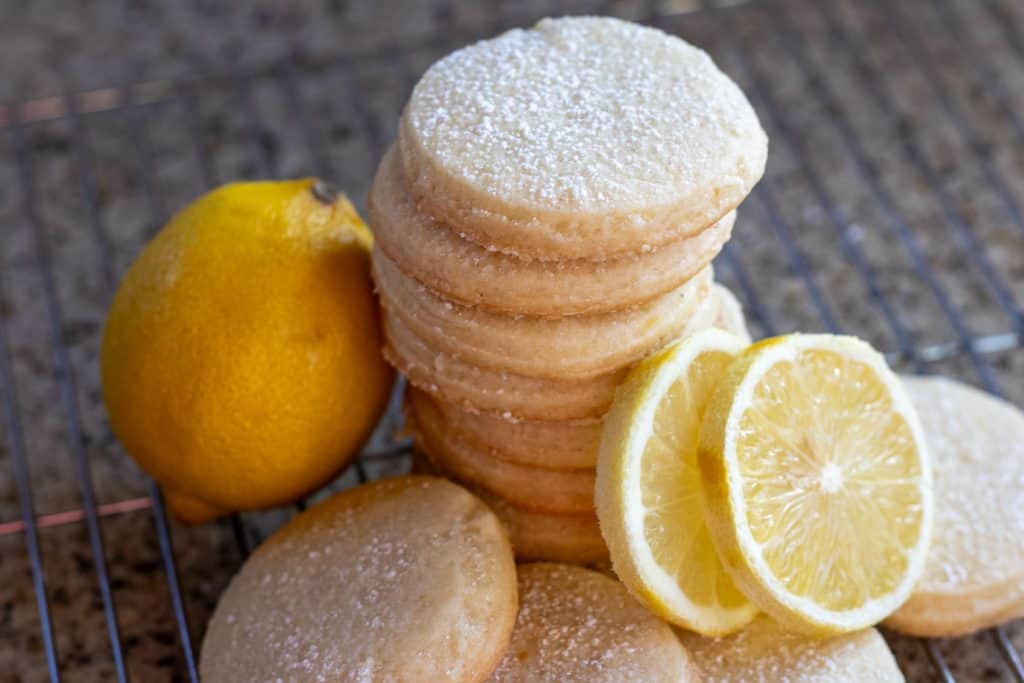 A stack of sourdough shortbread cookies dusted with powdered sugar and arranged next to a lemon and two slices of lemon rounds.
