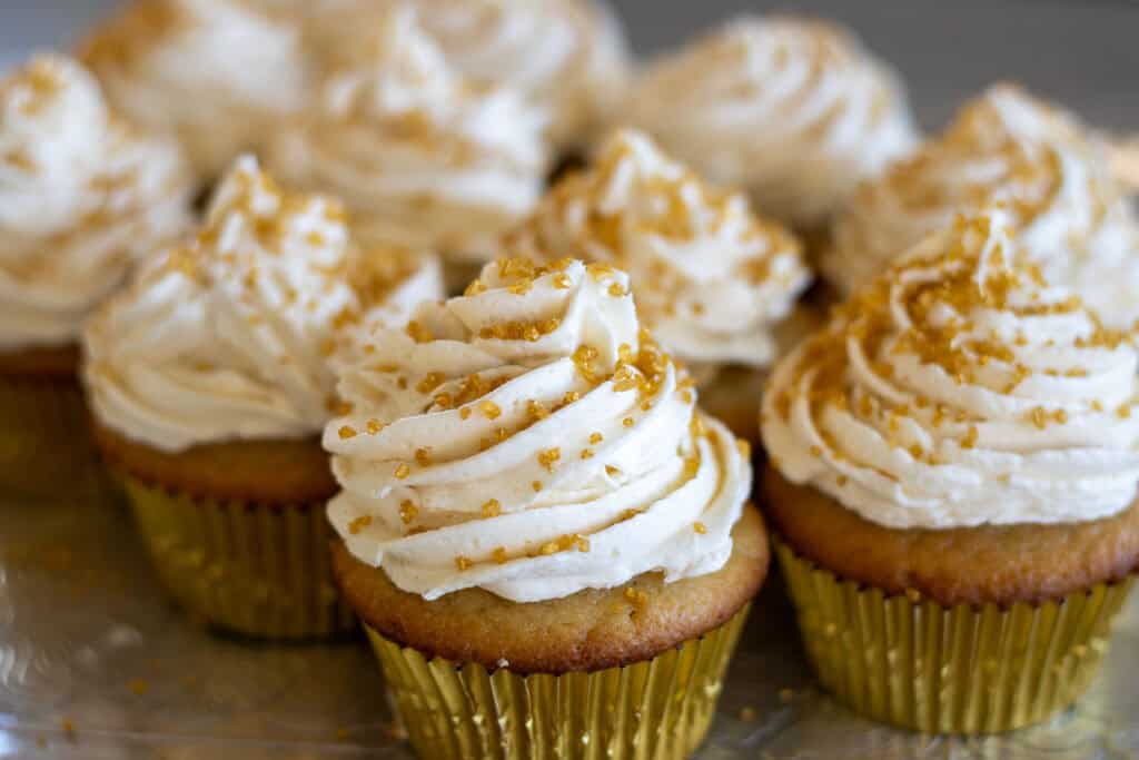 A batch of sourdough vanilla cupcakes with white buttercream frosting on top and golden sprinkles.