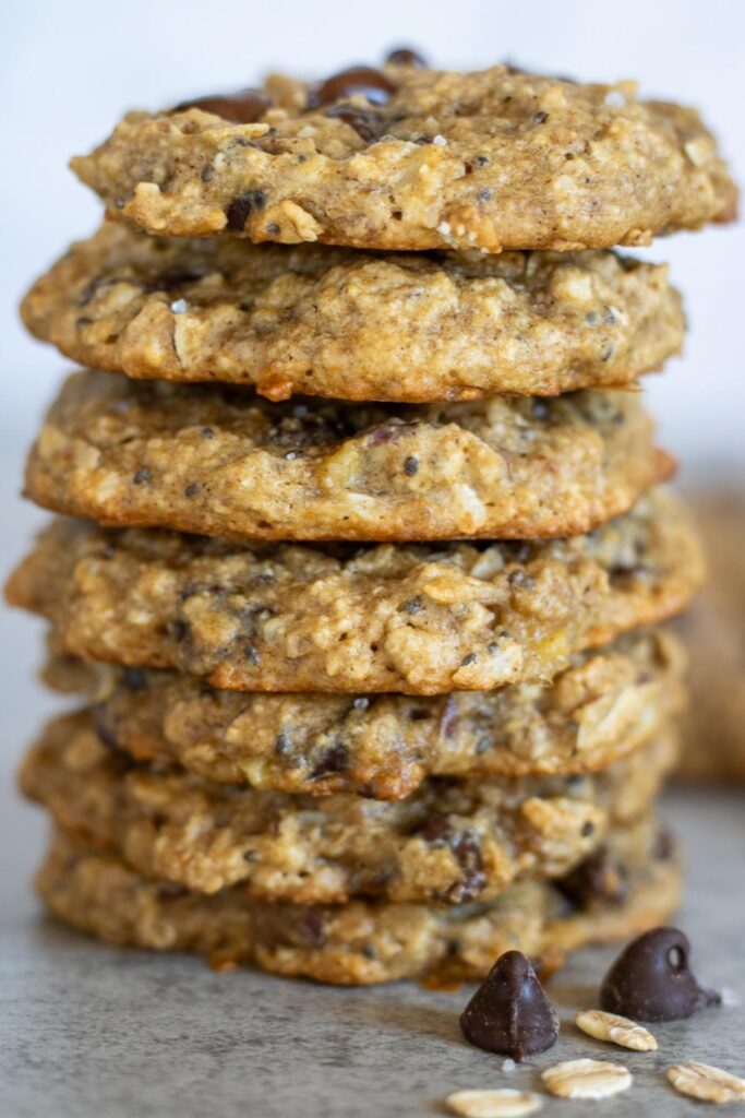 A stack of seven chocolate chip sourdough breakfast cookies with some chocolate chips and rolled oats.