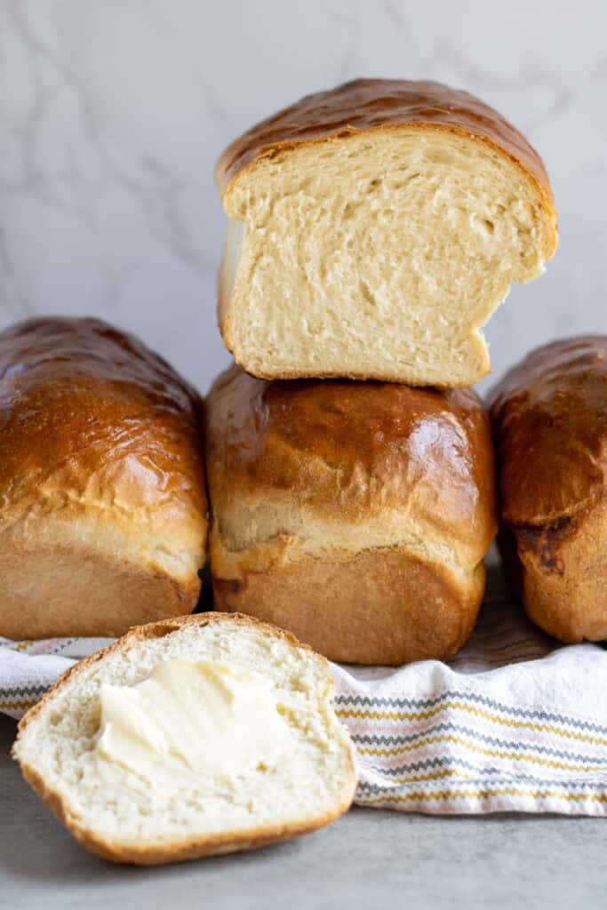 Three loaves of white sandwich bread on a platter with a sliced loaf resting on top of the middle loaf and one slice in the front with butter on it.