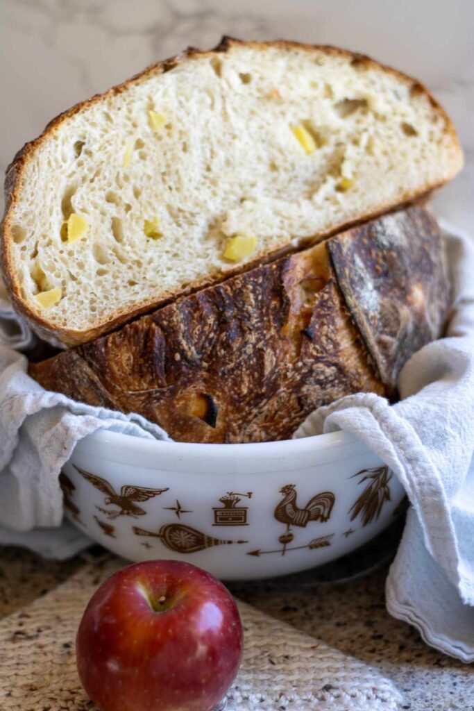 A bowl lined with a tea towel with a cut in half loaf of apple sourdough bread, with a whole apple in the foreground.