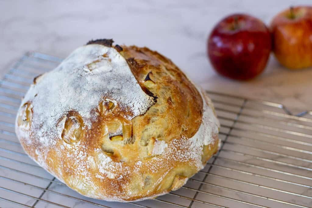 A sourdough apple bread boule cooling on a wire rack with two apples in the background.