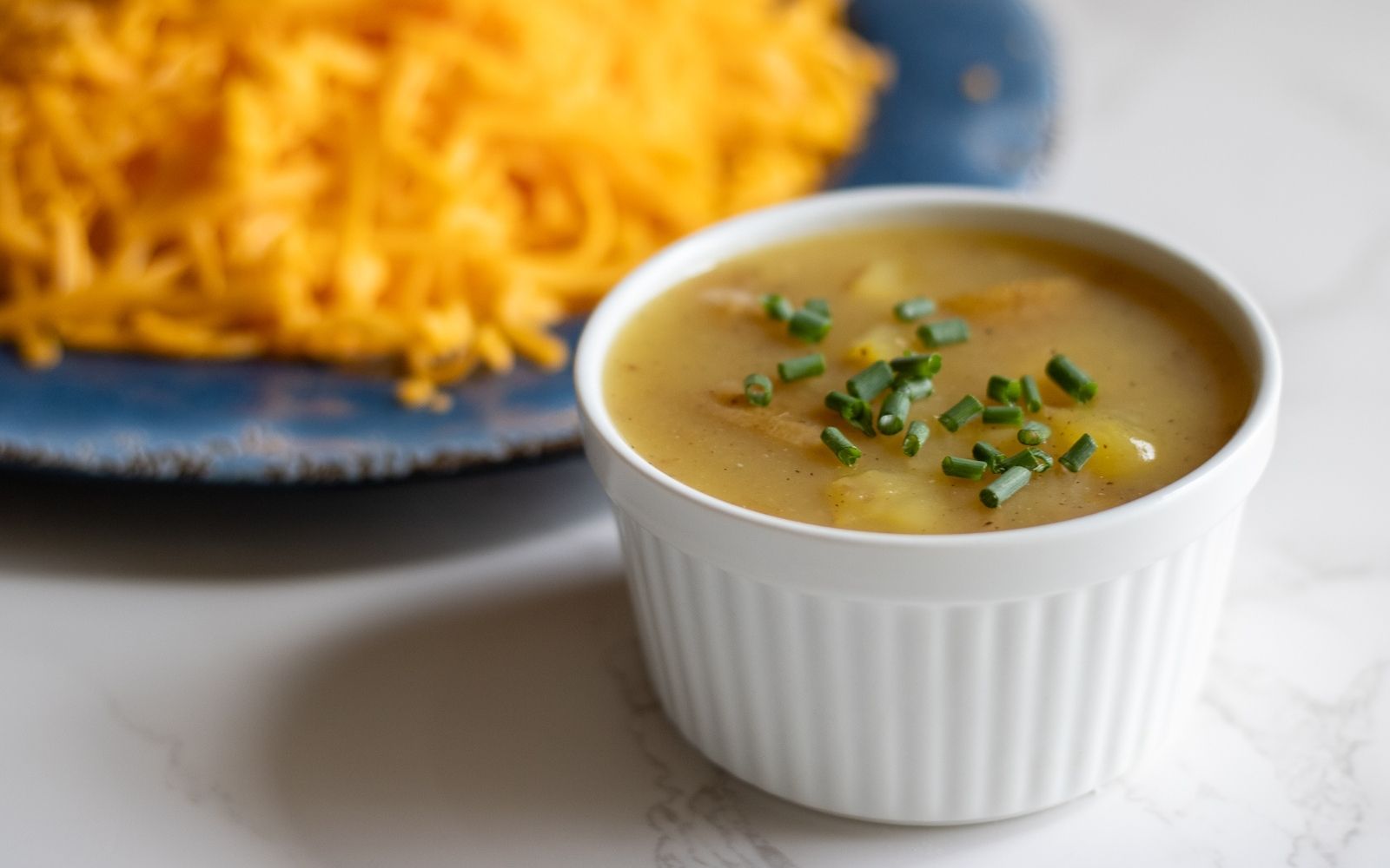 A white soup ramekin filled with 3 ingredient potato soup topped with chives with a plate full of shredded cheese.