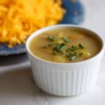 A small white bowl of 3 ingredient potato soup topped with fresh chives, served next to a plate of shredded cheddar cheese.