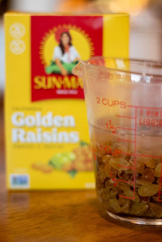 Golden raisins soaking in water inside a glass measuring cup, with a Sun-Maid Golden Raisins box in the background, preparing for sourdough hot cross buns.