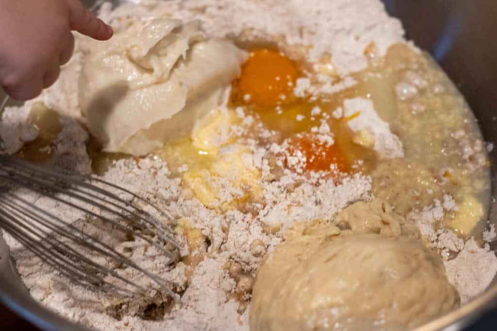 Child’s hand pointing at tangzhong in a mixing bowl filled with flour, eggs, sweet stiff starter, and other ingredients, ready to mix for sourdough hot cross buns.