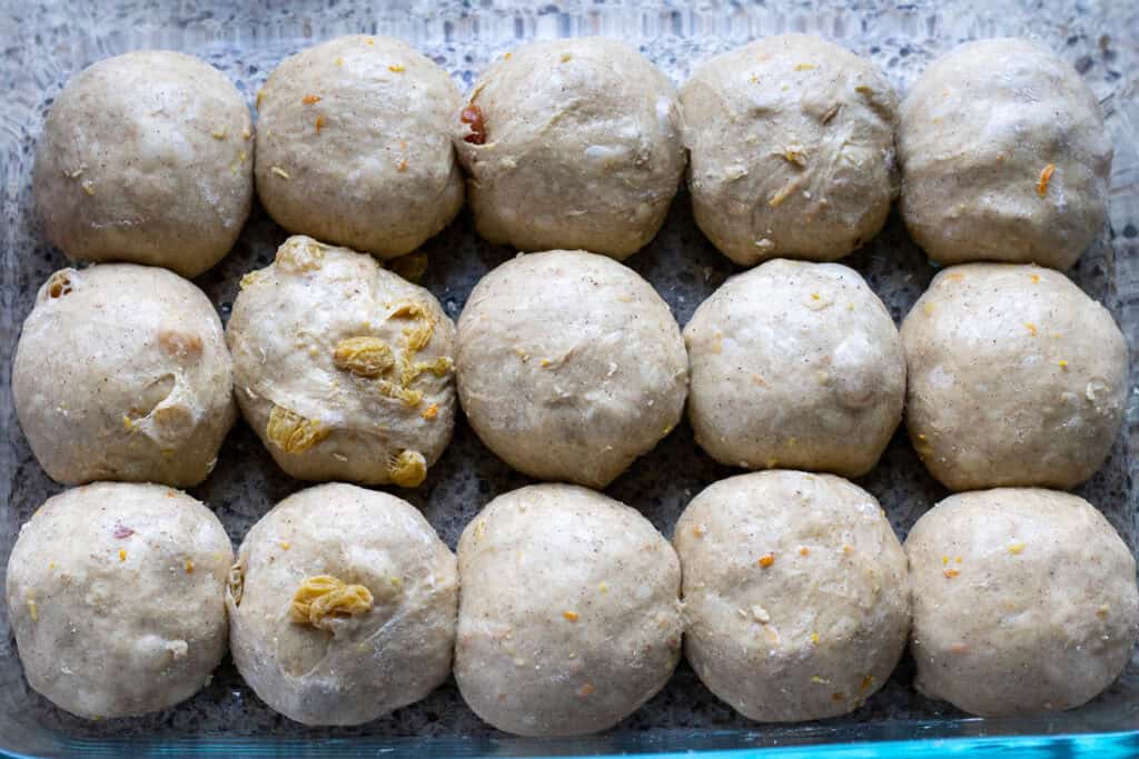 Fifteen shaped sourdough hot cross bun dough balls arranged in a greased glass baking dish, ready for final proofing.