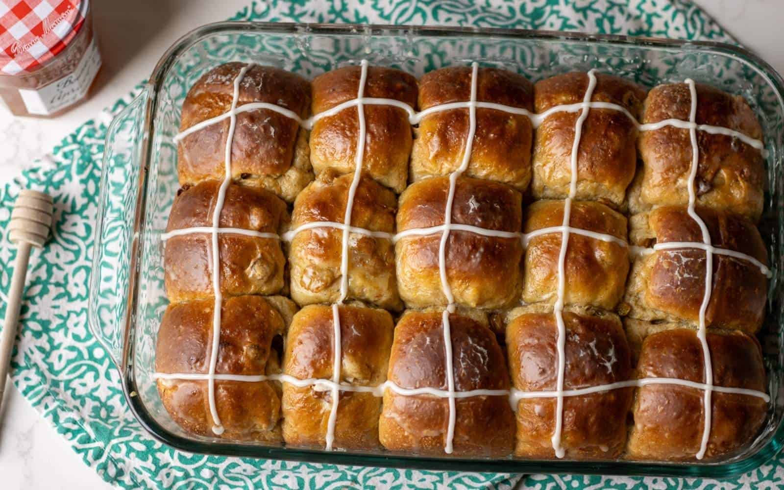 Freshly baked sourdough hot cross buns in a glass baking dish, topped with glossy orange glaze and sweet white icing crosses, with a jar of apricot jam and honey dipper on the side.