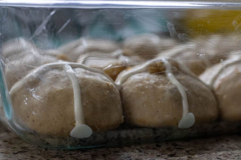 Unrisen hot cross buns with cross paste piped on top, covered in plastic wrap for final proofing.