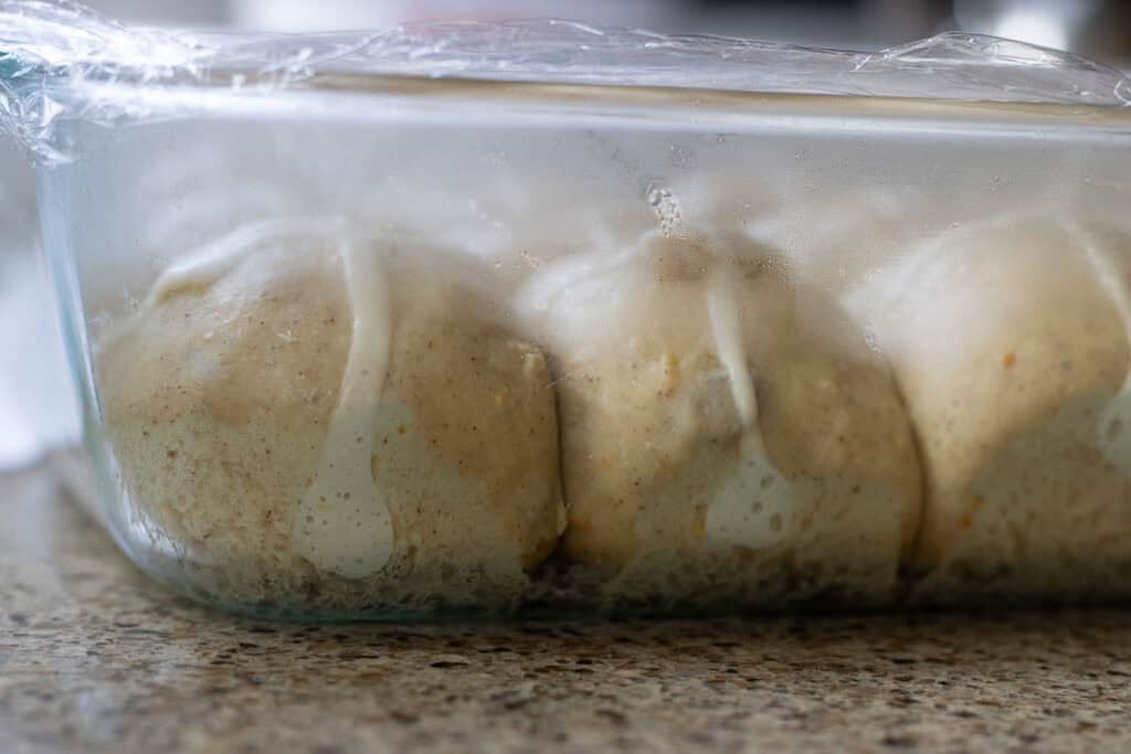 Close-up side view of proofed hot cross buns in a glass baking dish, paste for crosses visible under plastic wrap.