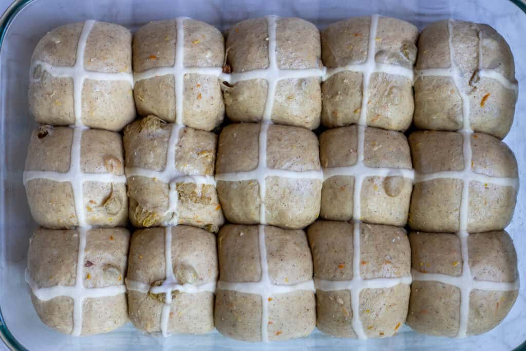 Risen sourdough hot cross buns in a glass baking dish, with piped flour and water paste crosses before baking.