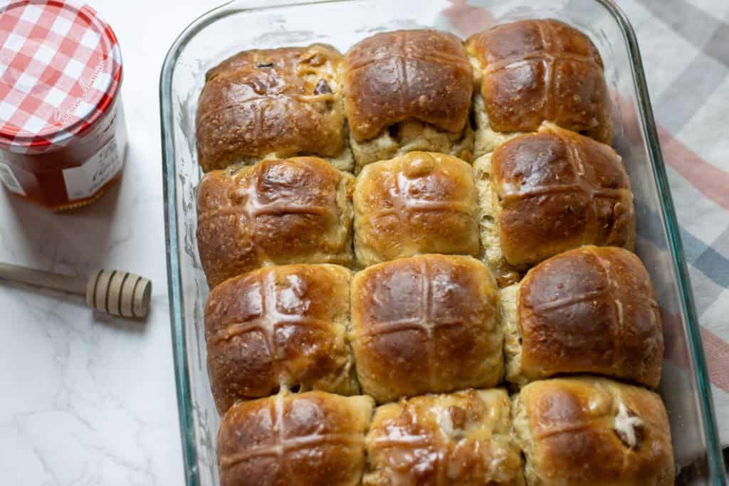 Glazed sourdough hot cross buns in a glass baking dish, brushed with orange glaze for a glossy finish, with a jar of Bonne Maman marmalade and honey dipper beside.