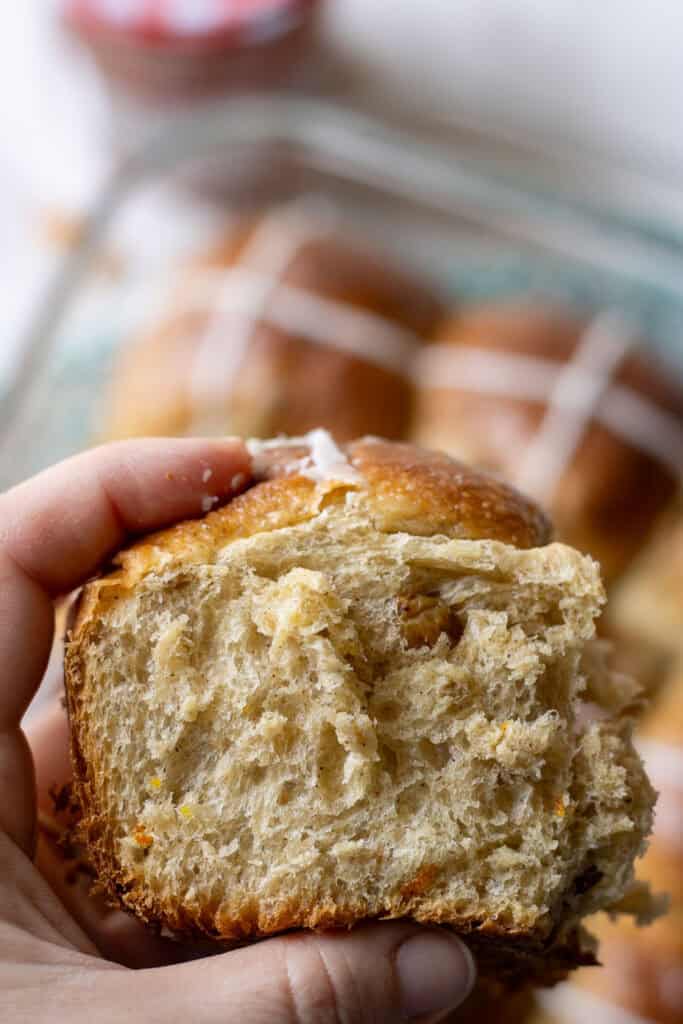 Close-up of a hand holding a fluffy, torn sourdough hot cross bun with golden raisins and a soft crumb, showing the texture of this tangzhong sourdough hot cross buns recipe.