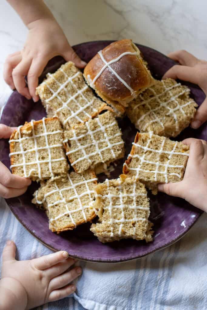 Children’s hands reaching for sliced sourdough hot cross buns with icing, arranged on a purple plate over a striped cloth, showcasing the soft texture and playful family moment.
