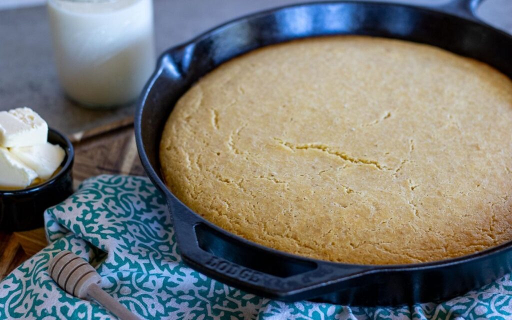 Whole sourdough cornbread baked in a cast iron skillet, with butter, honey dipper, and a jar of milk in the background.