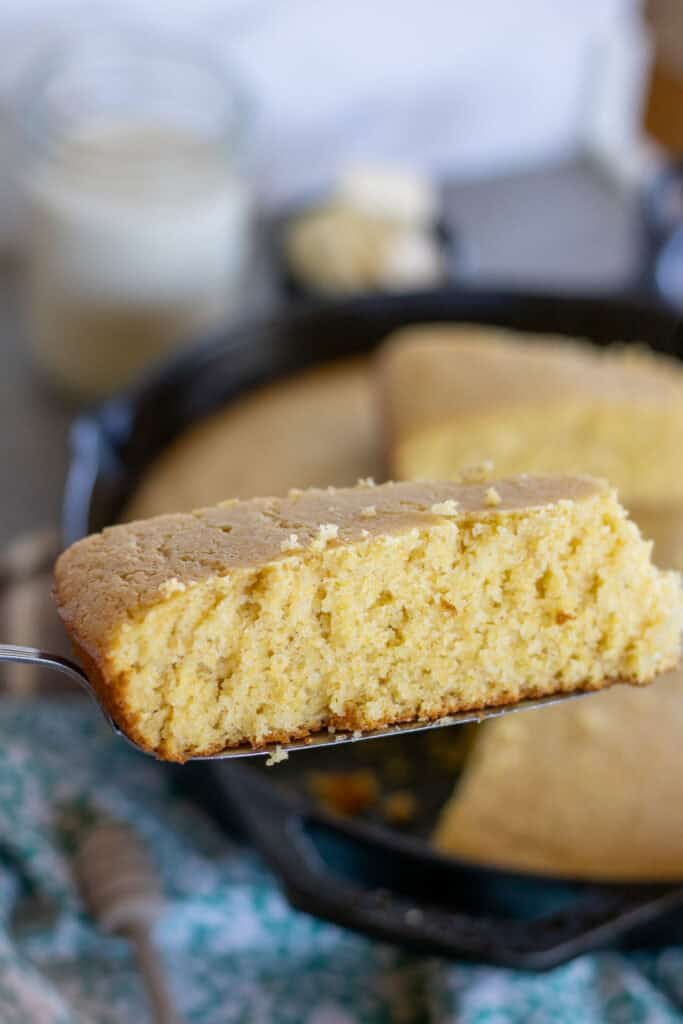 A slice of golden sourdough cornbread held over a cast iron skillet, showing its moist, tender crumb.