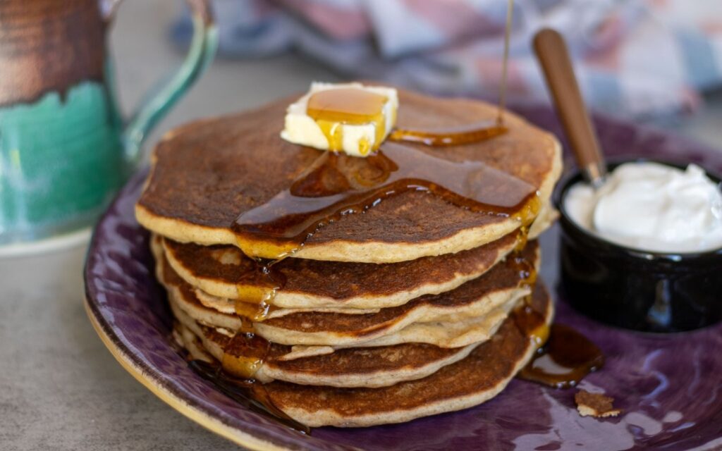 Stack of sourdough cottage cheese pancakes topped with butter and syrup, served on a purple plate with syrup dripping down the sides and a small bowl of plain yogurt on the side