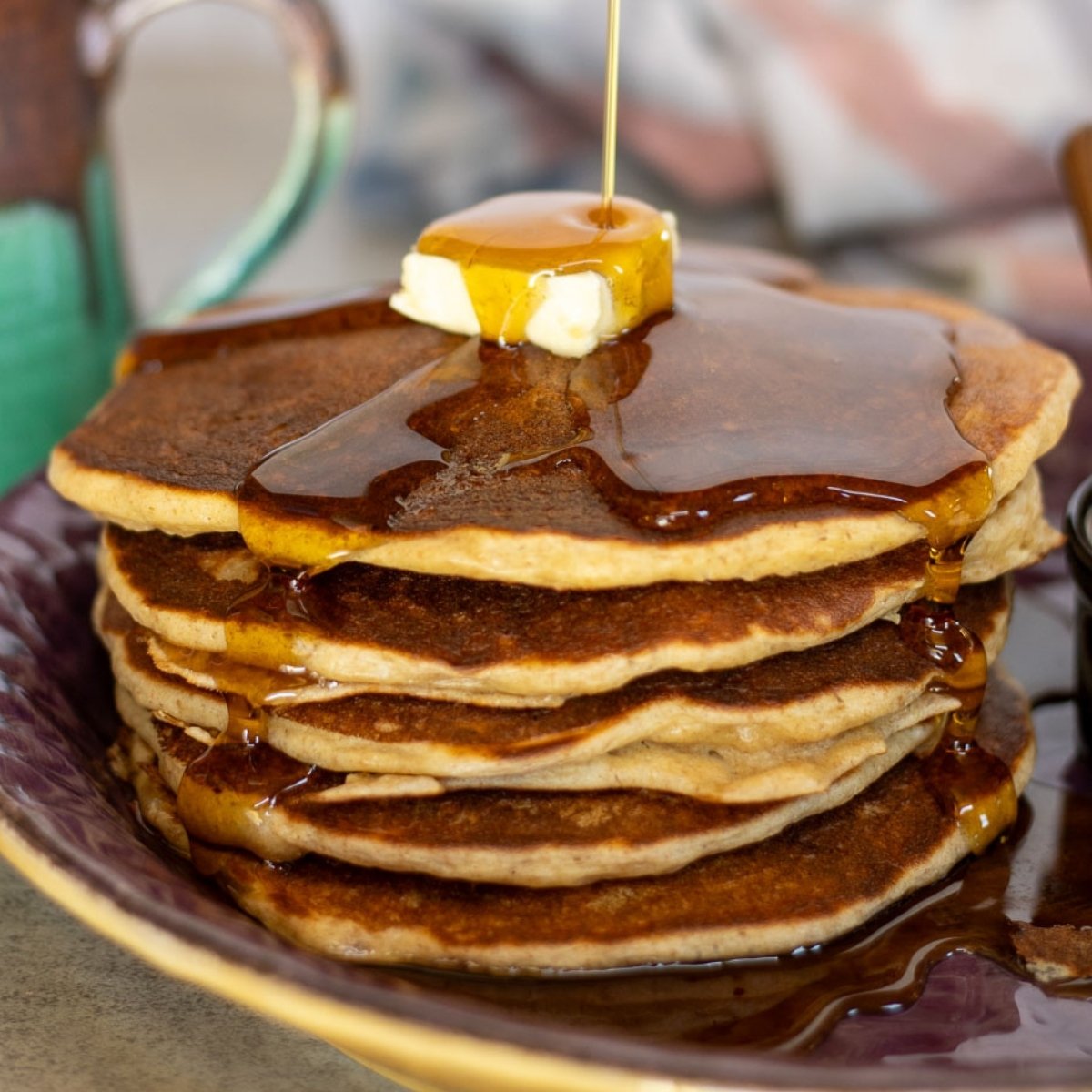 Stack of golden-brown sourdough cottage cheese pancakes topped with a pat of butter and maple syrup being drizzled over the top, served on a deep purple plate