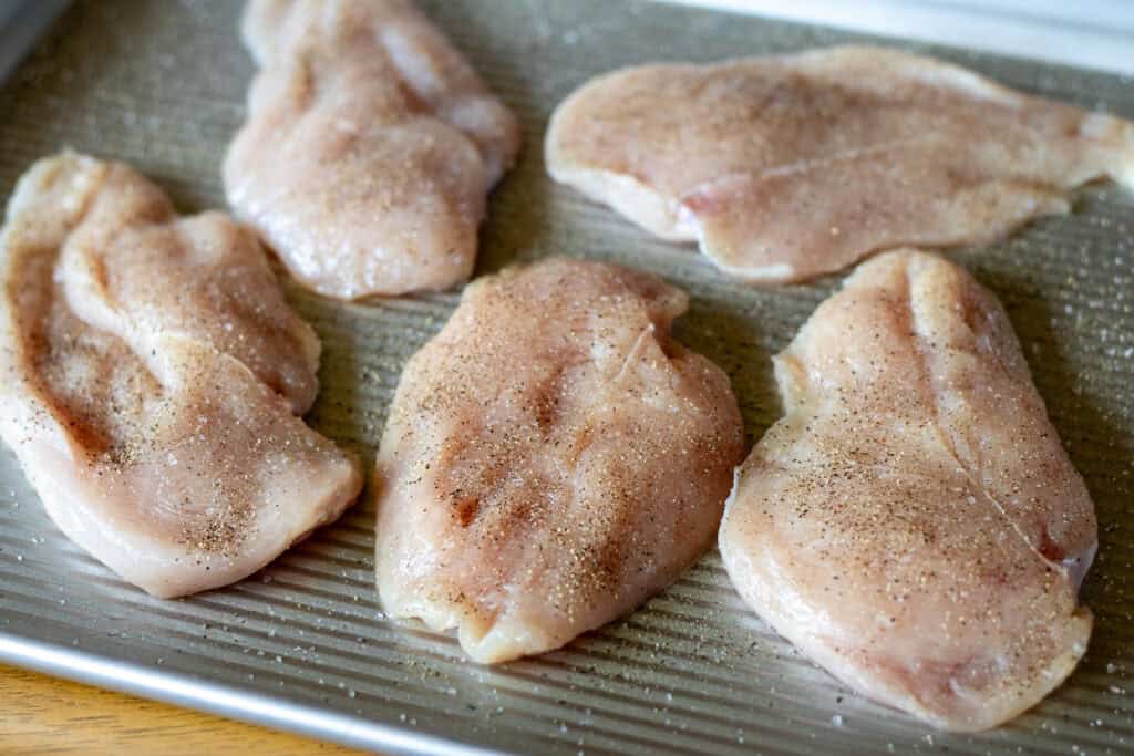 "Small boneless skinless chicken breasts seasoned with salt and pepper on a baking sheet, ready to be seared.