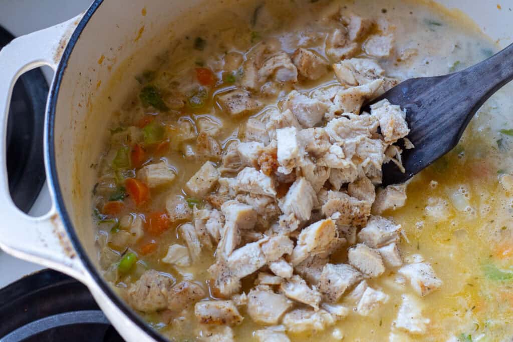 Diced cooked chicken being stirred into a pot of homemade gravy with carrots and celery