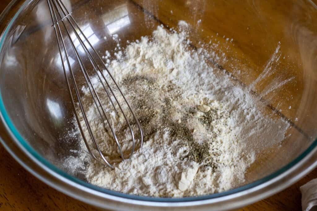 Glass mixing bowl filled with flour and dried herbs, ready to be whisked for dumpling dough.