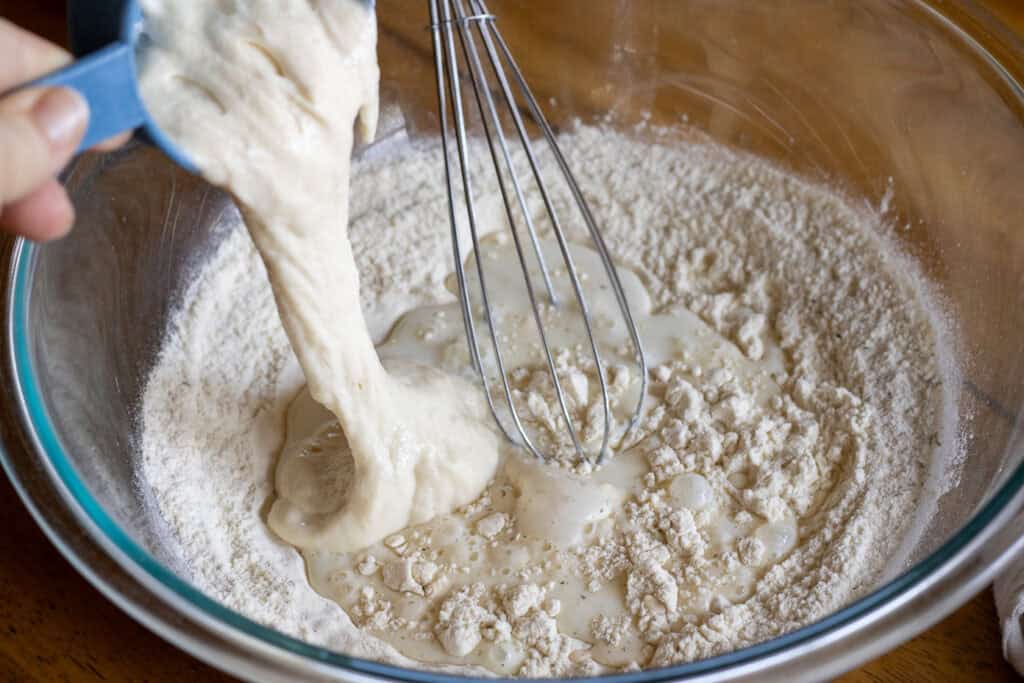 Sourdough starter being poured into a glass mixing bowl of flour and milk with a whisk, starting the dough for homemade dumplings.