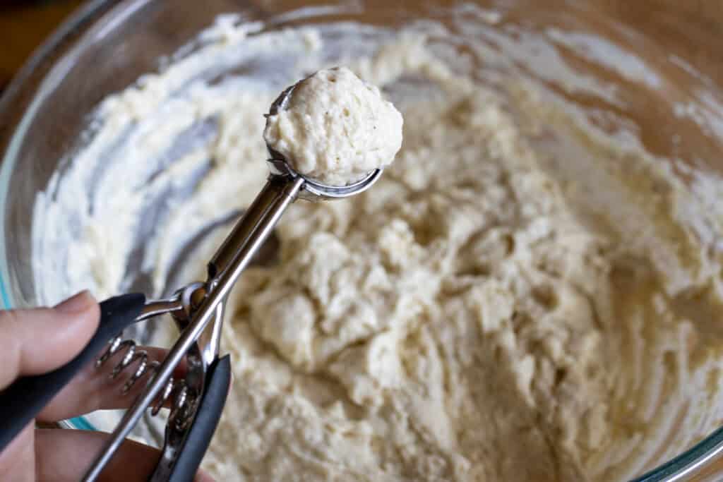 A small cookie scoop filled with sticky dumpling dough held above a glass bowl of mixed batter, ready to be dropped into simmering soup.