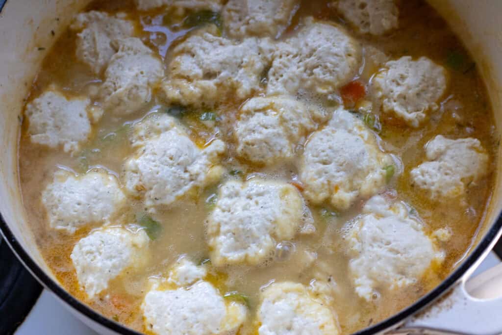 Uncooked sourdough dumplings just added to bubbling chicken gravy in a white Dutch oven, ready to be covered and steamed.