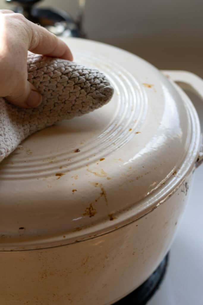 A hand holding a pot holder presses down the lid of a white Dutch oven, sealing in steam while dumplings cook inside.