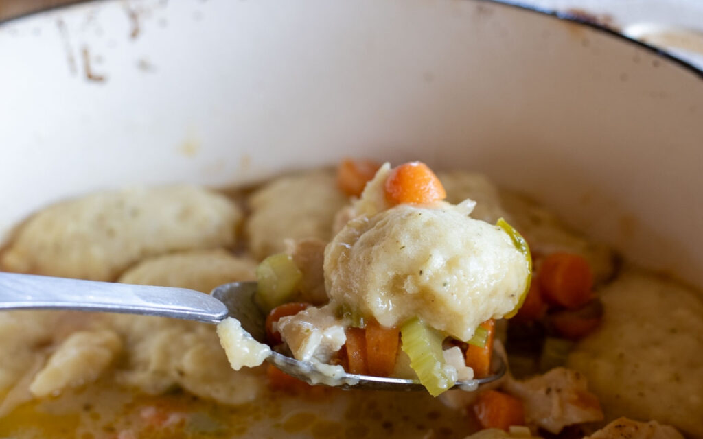 Close-up of a spoonful of sourdough dumpling with carrots, celery, and chicken lifted from a Dutch oven filled with bubbling homemade chicken and dumplings.