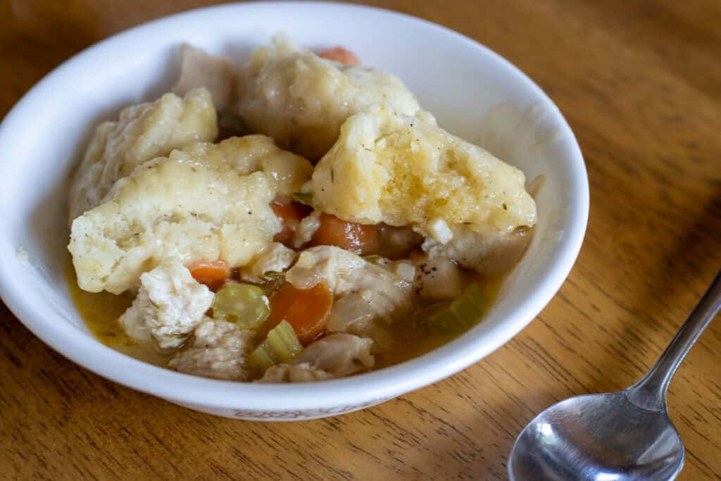 A white bowl filled with homemade discard dumplings, carrots, celery, and chicken in a savory broth, served on a wooden table with a spoon nearby.