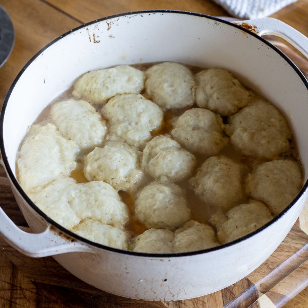 Overhead view of freshly steamed sourdough dumplings in a white Dutch oven, showing fluffy, biscuit-like tops cooked in bubbling broth on a wooden cutting board.