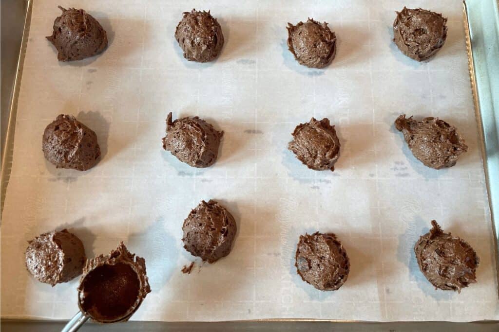 A baking sheet with sourdough double chocolate cookie batter balls spaced about two inches apart with a cookie scoop showing the sticky texture of the batter.